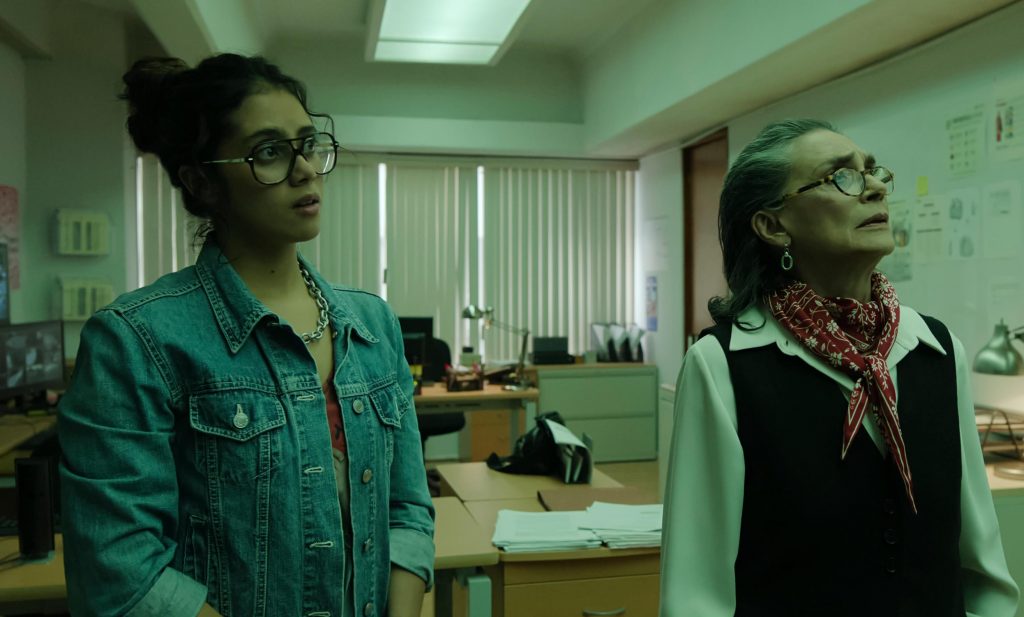 Dans un bureau universitaire, 2 femmes sont de face, légèrement 3/4 et regardent vers le haut à droite. Celle de gauche est une jeune femme, les cheveux en chignon avec une veste en jean et des lunettes. Celle de droite est plutôt âgée, porte une chemise et un gilet ainsi qu'un foulard au cou, elle porte également des lunette. Elle a des cheveux court/mi-long détachés mais coiffé vers l'arrière.