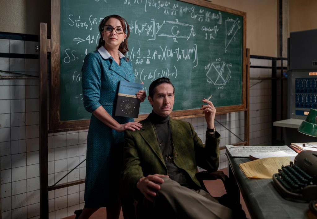 Devant un tableau rempli d'équations, un couple des années 60 nous regarde. La femme est debout légèrement derrière son mari. Elle porte un tailleur bleu et tient un livre de physique dans une main. L'autre est posé sur l'épaule de son mari. Celui-ci est assi dans un fauteuil. Il porte une veste verte, un col roulé noir. D'une main, il tient une cigarette. Les deux ont des lunette mais la femme les porte tandis que l'homme les a qui pendent à son cou.